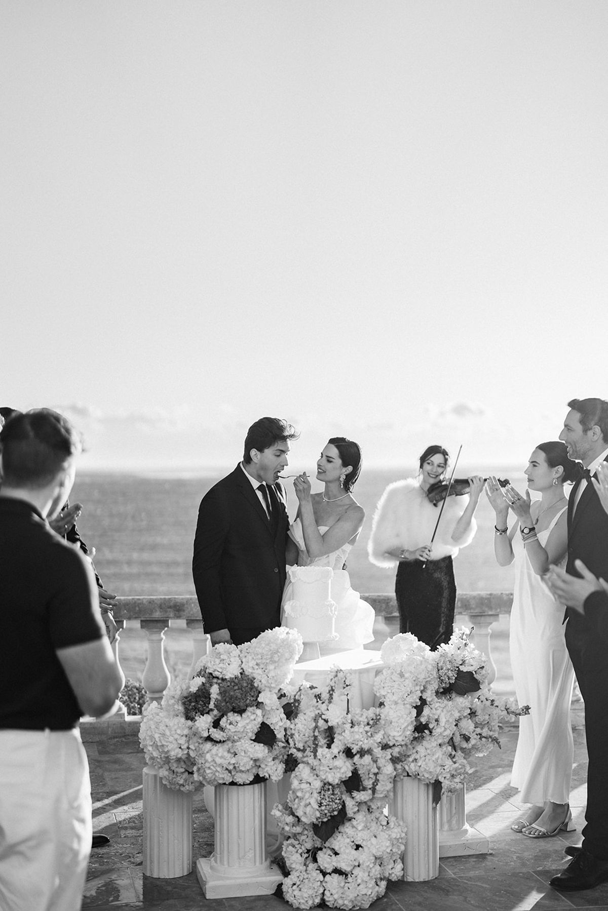 The groom feeds the bride a spoonful of cake as a violinist plays behind them and guests look on.