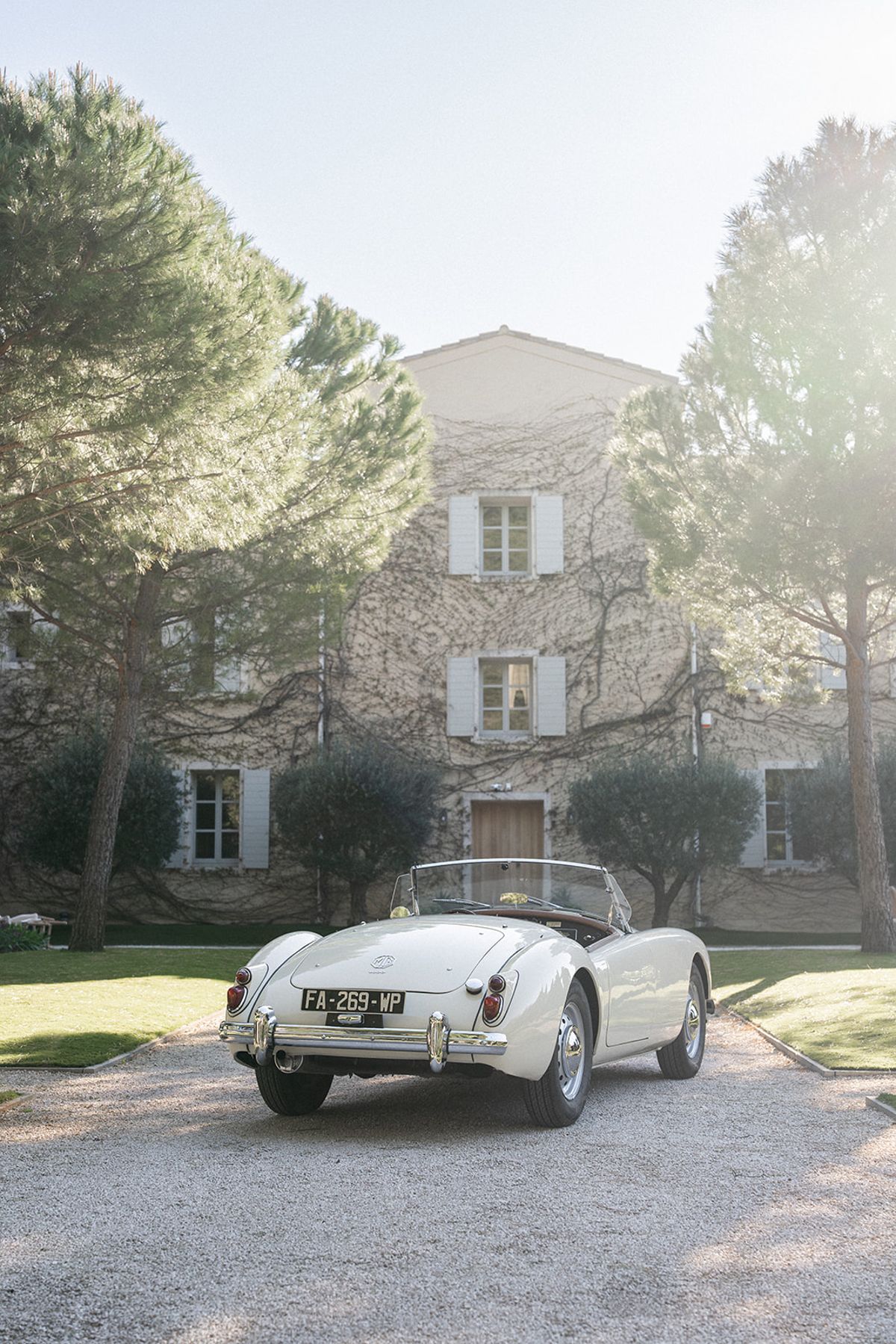A cream vintage MG convertible is parked in the gravel drive in front of the ivy-covered estate.