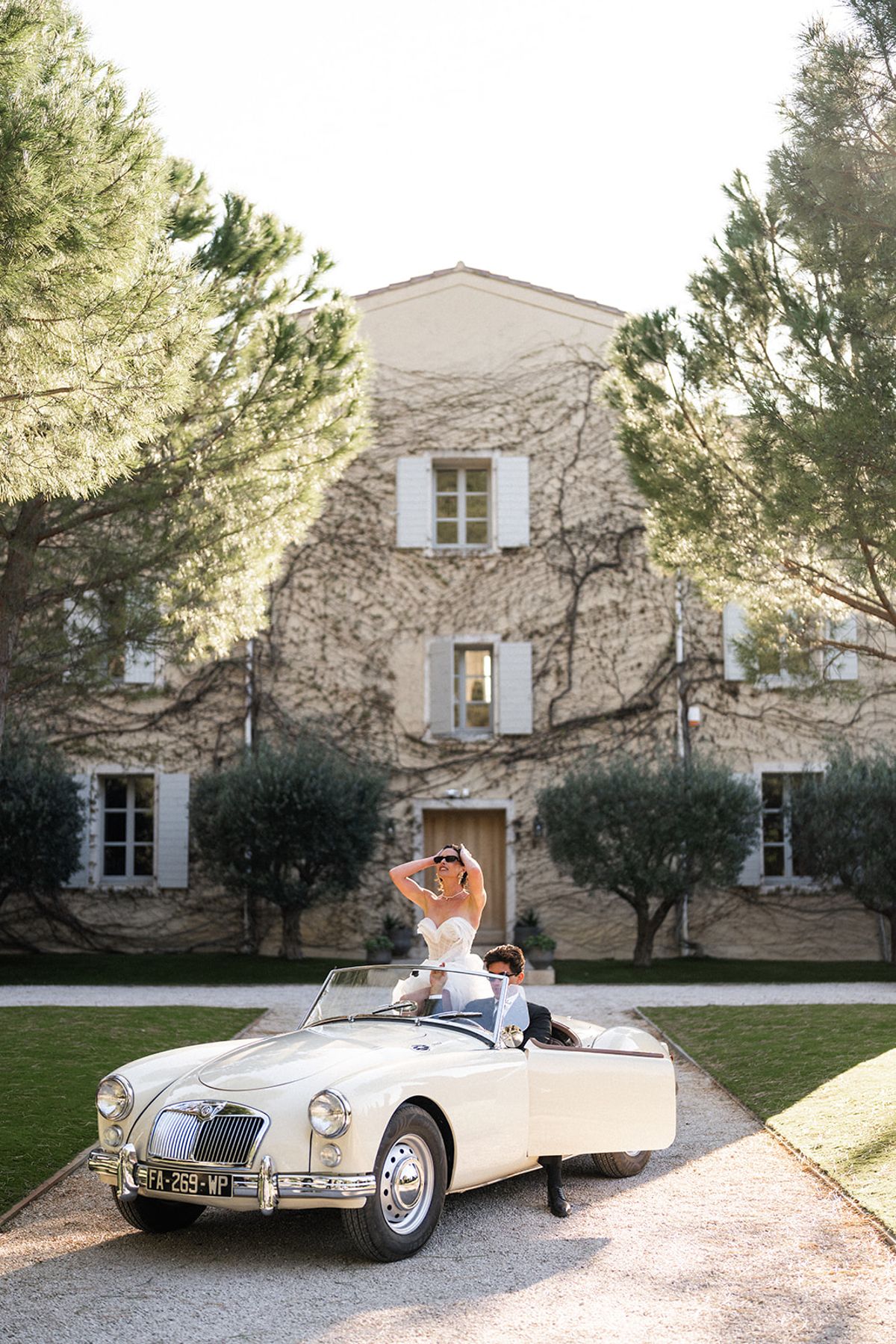 The bride and groom ride away from the estate in a vintage convertible, she laughing with a hand to her sunglasses.