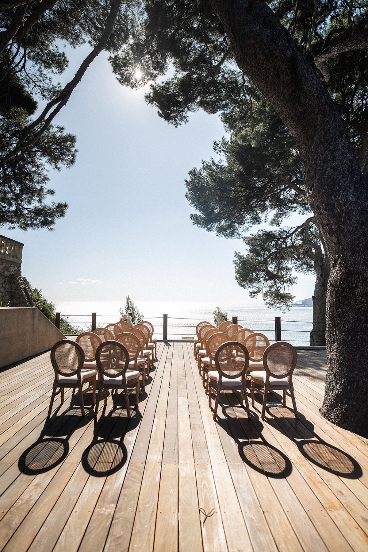 Rows of cane-back chairs stand empty on the wooden ceremony deck, facing the open sea beneath pine trees.