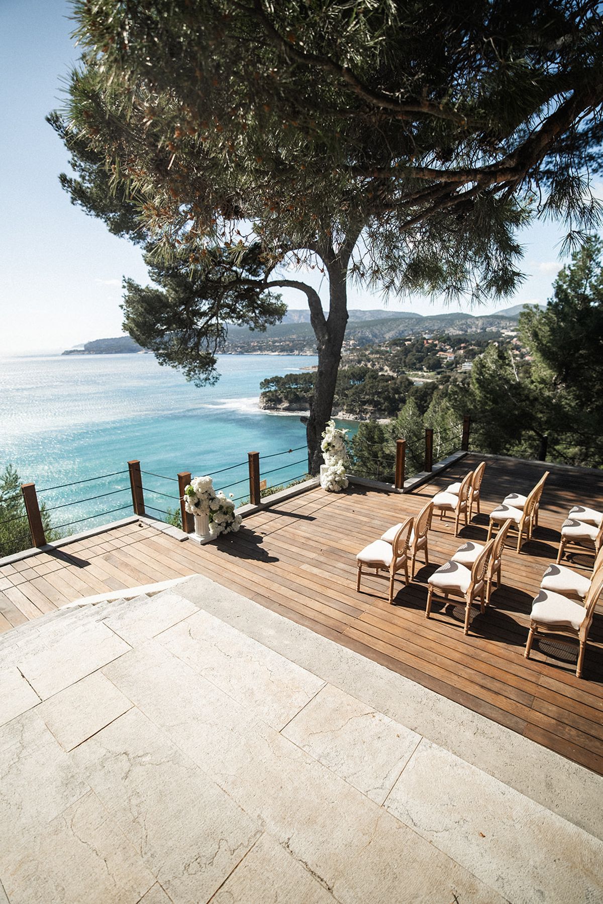 Ceremony seating set beneath pine trees on the cliffside deck, the sea framed between the branches.