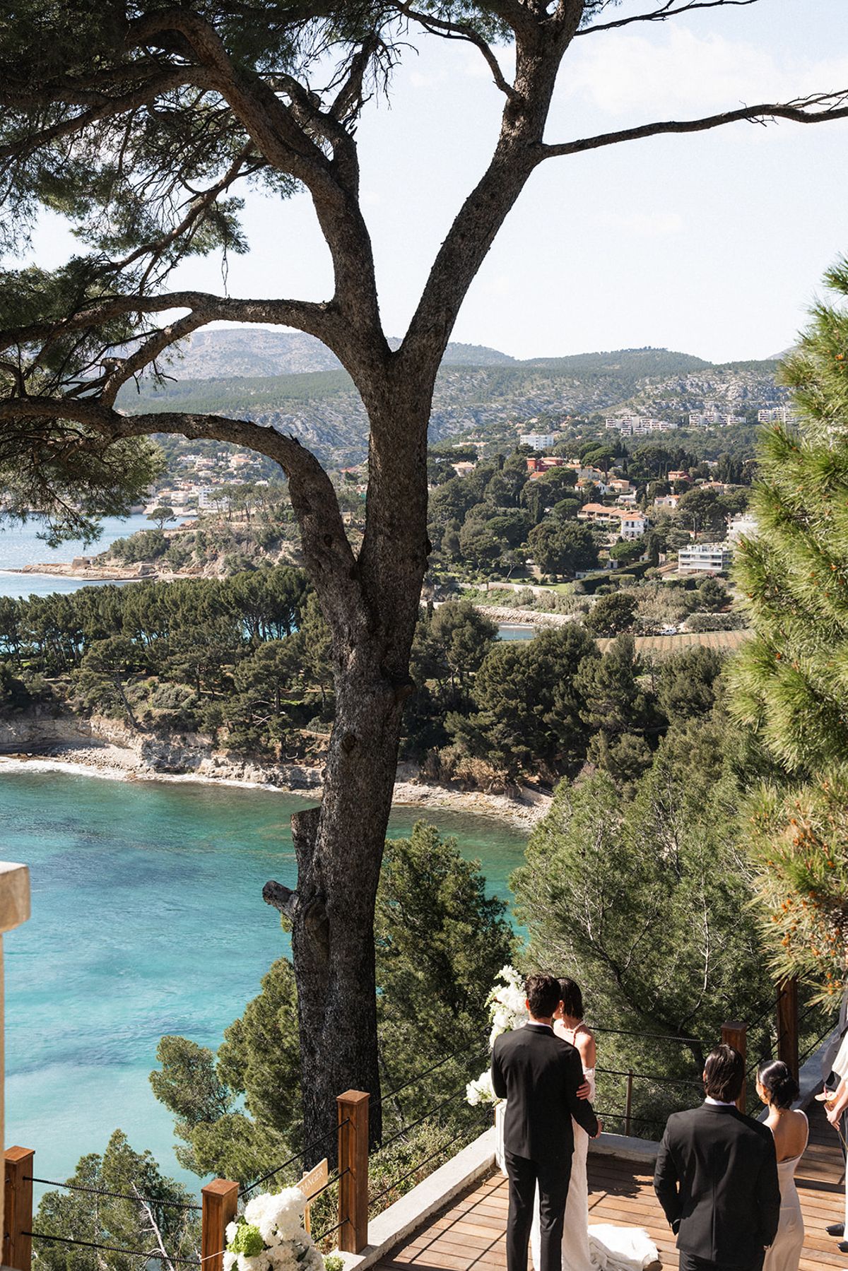 The bride crosses the ceremony deck past standing guests, the sea behind her.