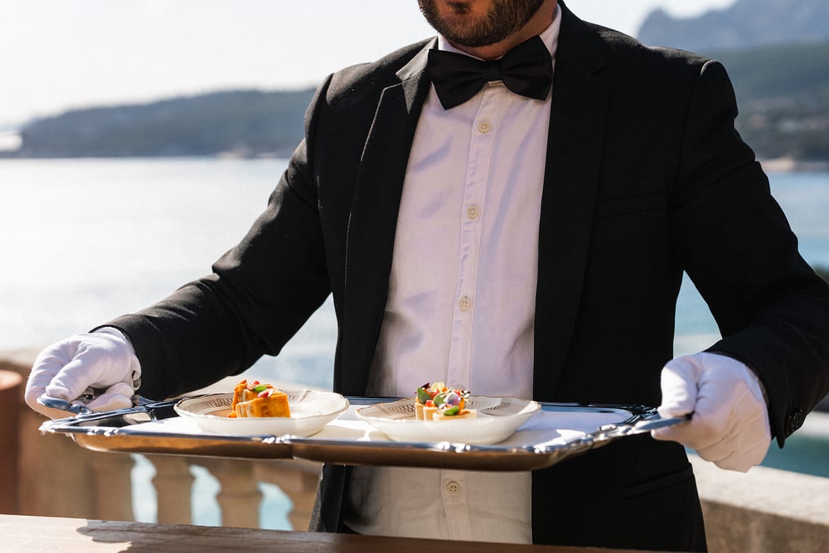 A white-gloved waiter presents a tray of two canapés, the coastline visible behind him.