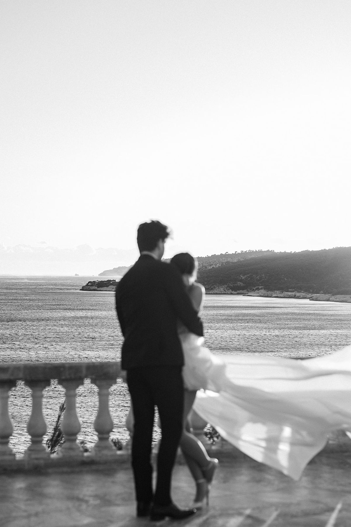Bride and groom embrace on a stone terrace overlooking the Mediterranean at sunset, her dress caught mid-movement in the wind.