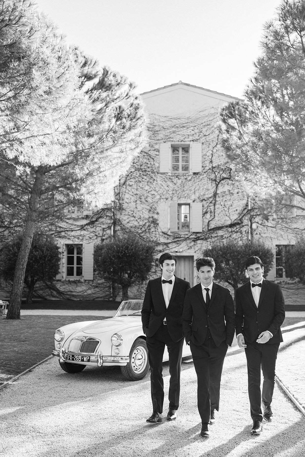 Three groomsmen in black tie walk up the gravel drive past the vintage car, the ivy-covered estate behind them.