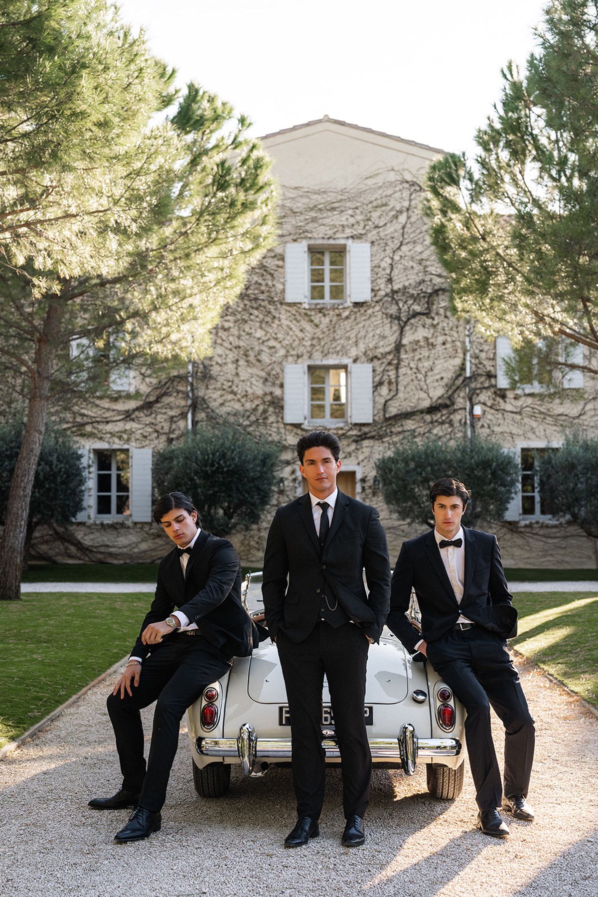 Three groomsmen in black tie stand together at a glass balustrade, pine trees visible through the window behind them.