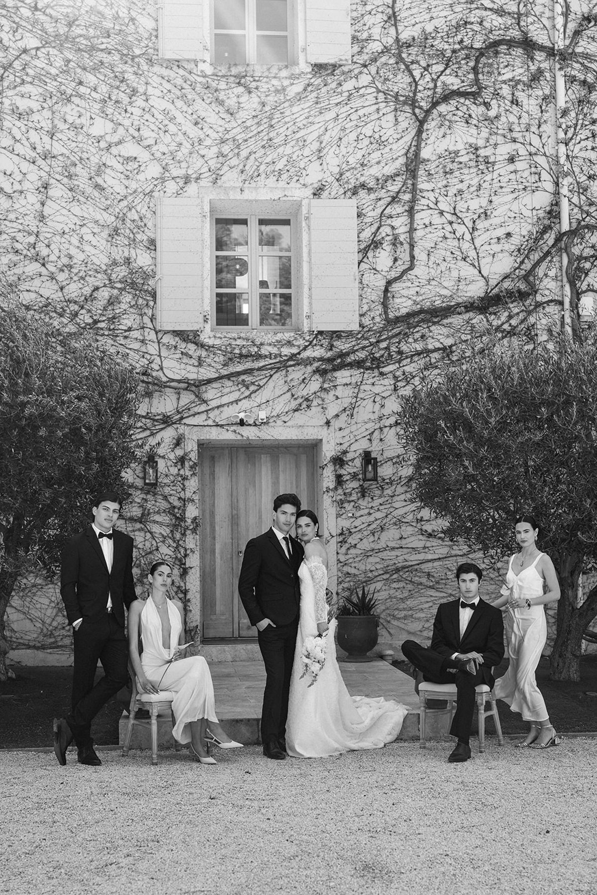 Bride and groom stand at the estate entrance with four members of the wedding party seated and standing around them, the ivy-covered facade behind.
