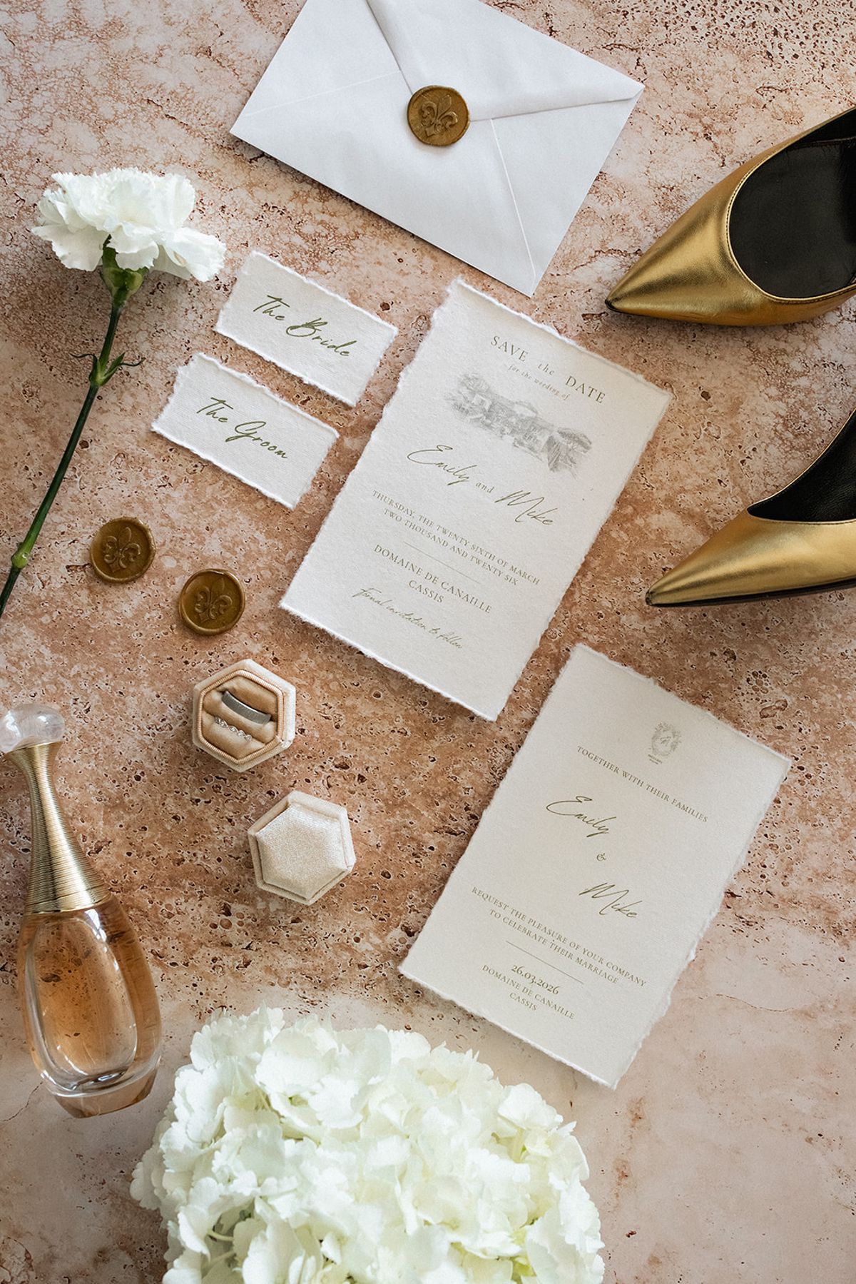 A flat lay of the save-the-date and invitation cards beside a ring box, perfume bottle, wax seals and gold heels.