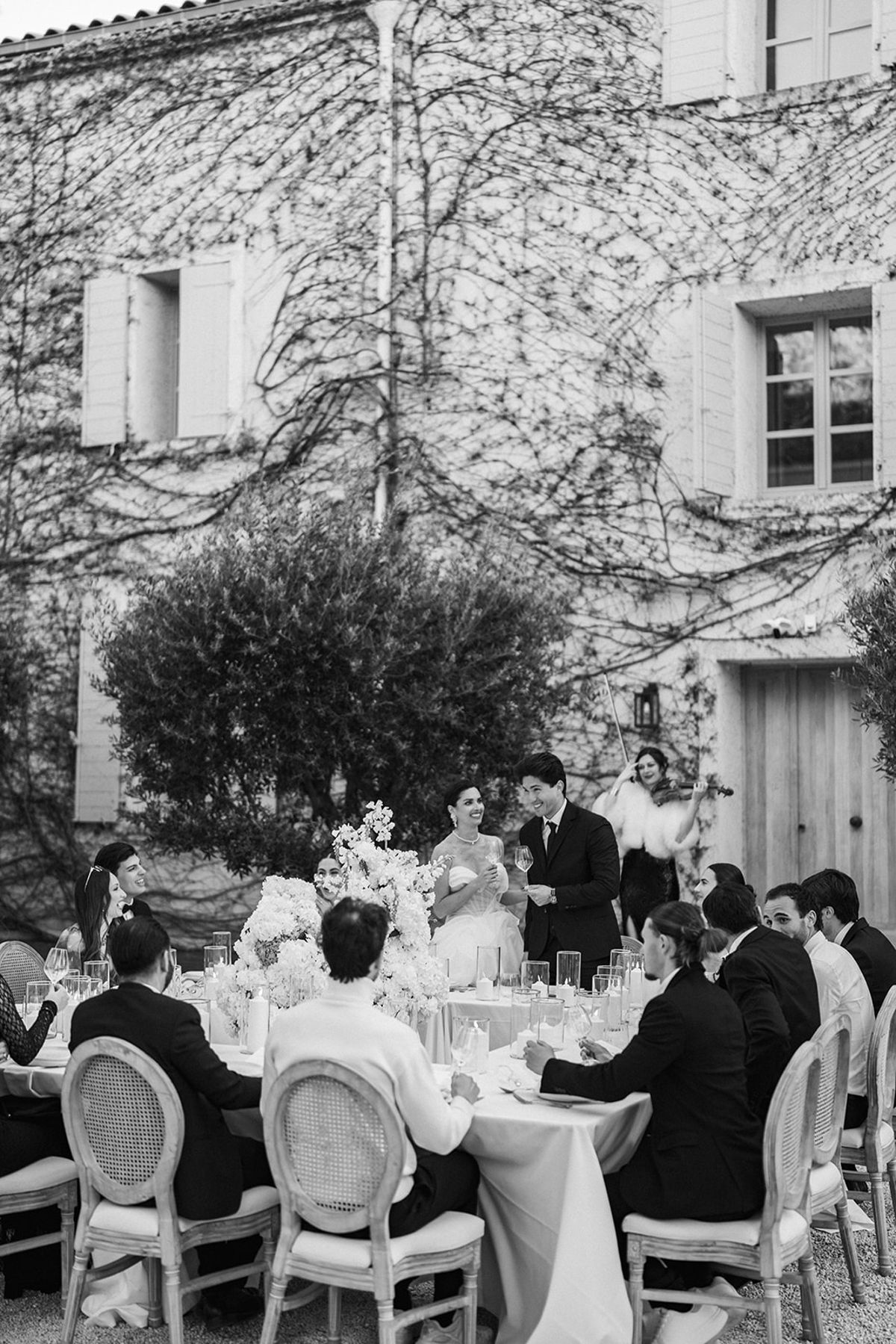 The bride and groom stand at the head of the reception table raising a toast as a violinist plays and guests applaud.