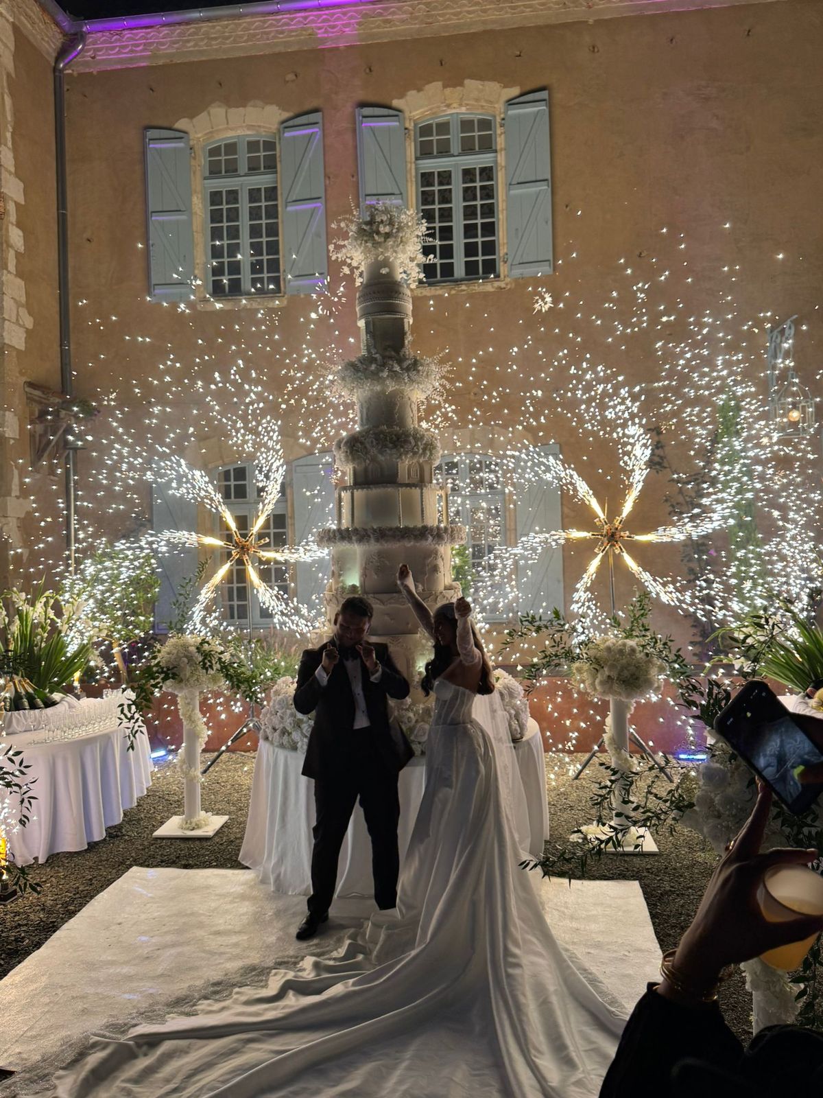 A couple cutting their wedding cake in the courtyard at night, surrounded by cold-spark fireworks.