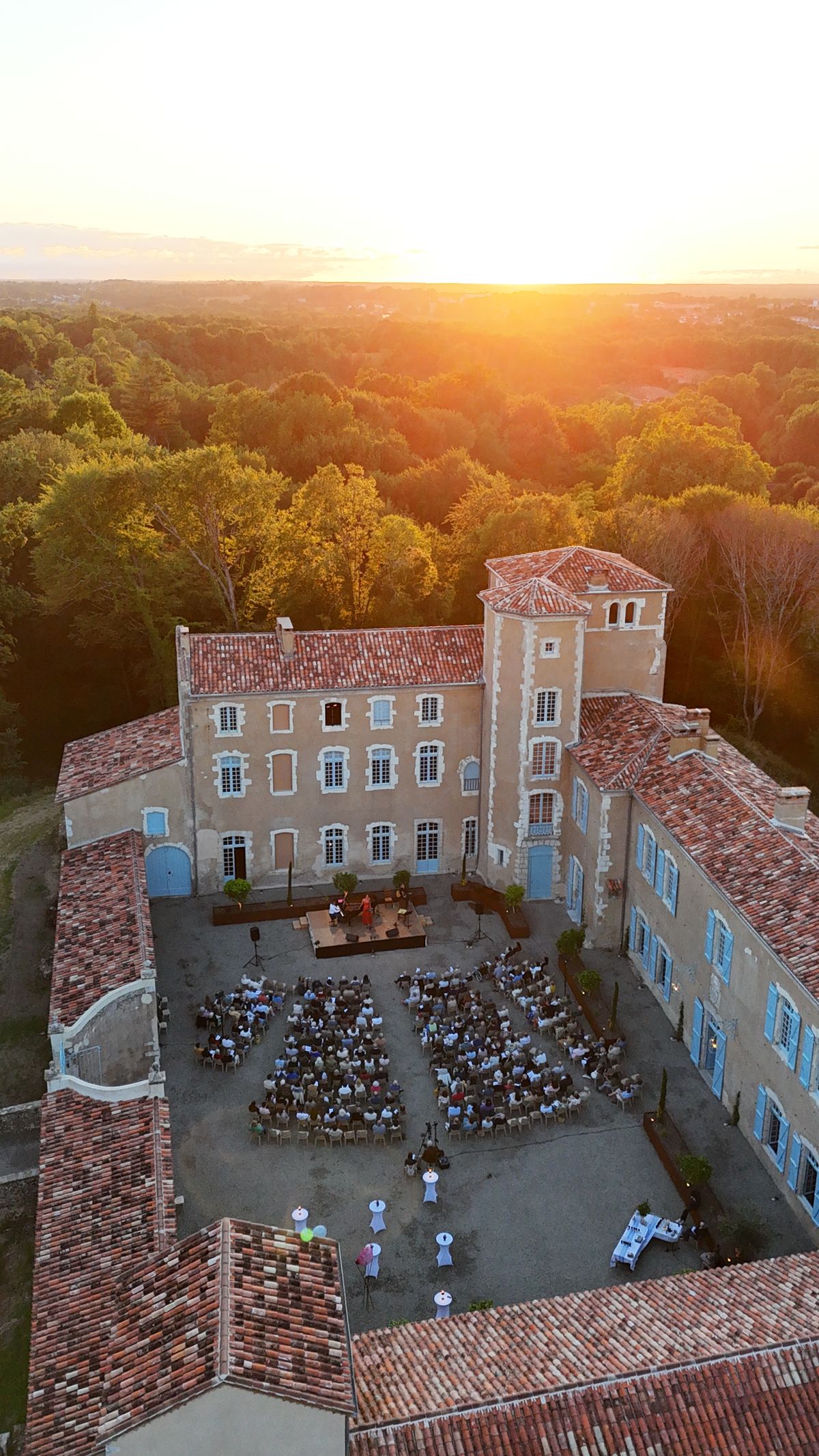 Aerial view at sunset of guests seated in rows in the château courtyard facing a stage, with the surrounding forest beyond.