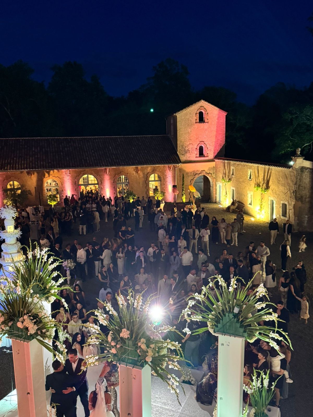 Guests gathered at night in the château courtyard for a wedding reception, the building lit in pink uplighting, tall floral arrangements in the foreground.