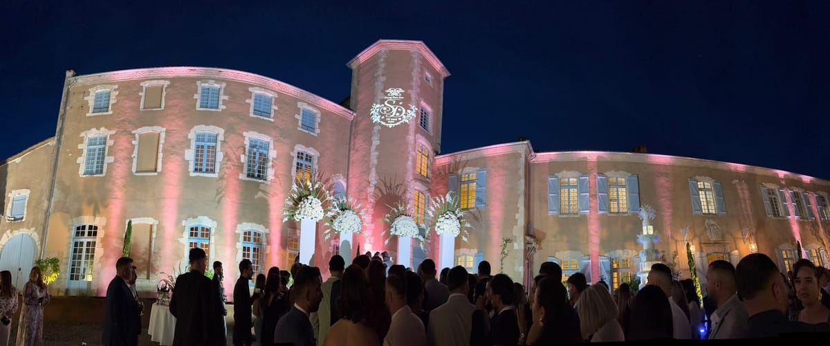 A wide view of guests gathered at night in the château courtyard for a reception, the building lit in pink.