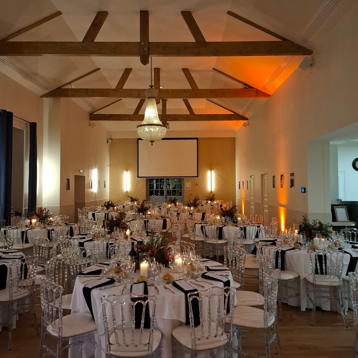 A reception hall set for a formal dinner with clear chairs, black and white table settings and a beamed ceiling.