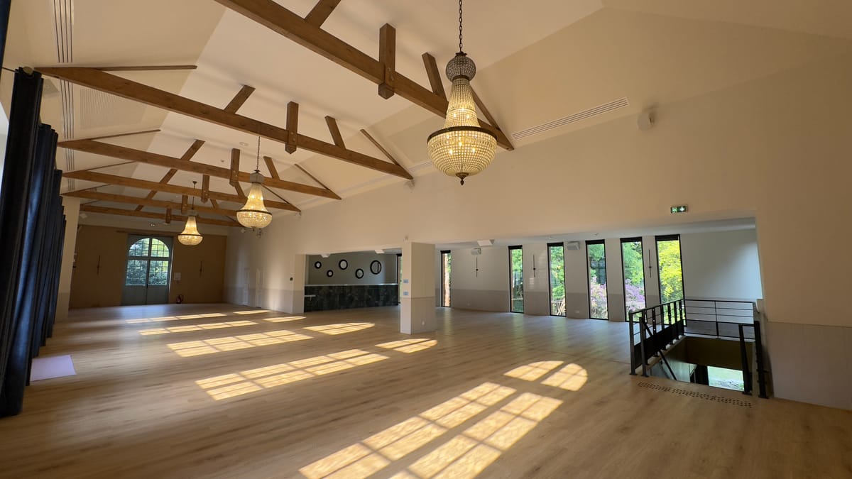 An empty reception hall with a beamed ceiling, chandeliers and glass doors opening onto the grounds.