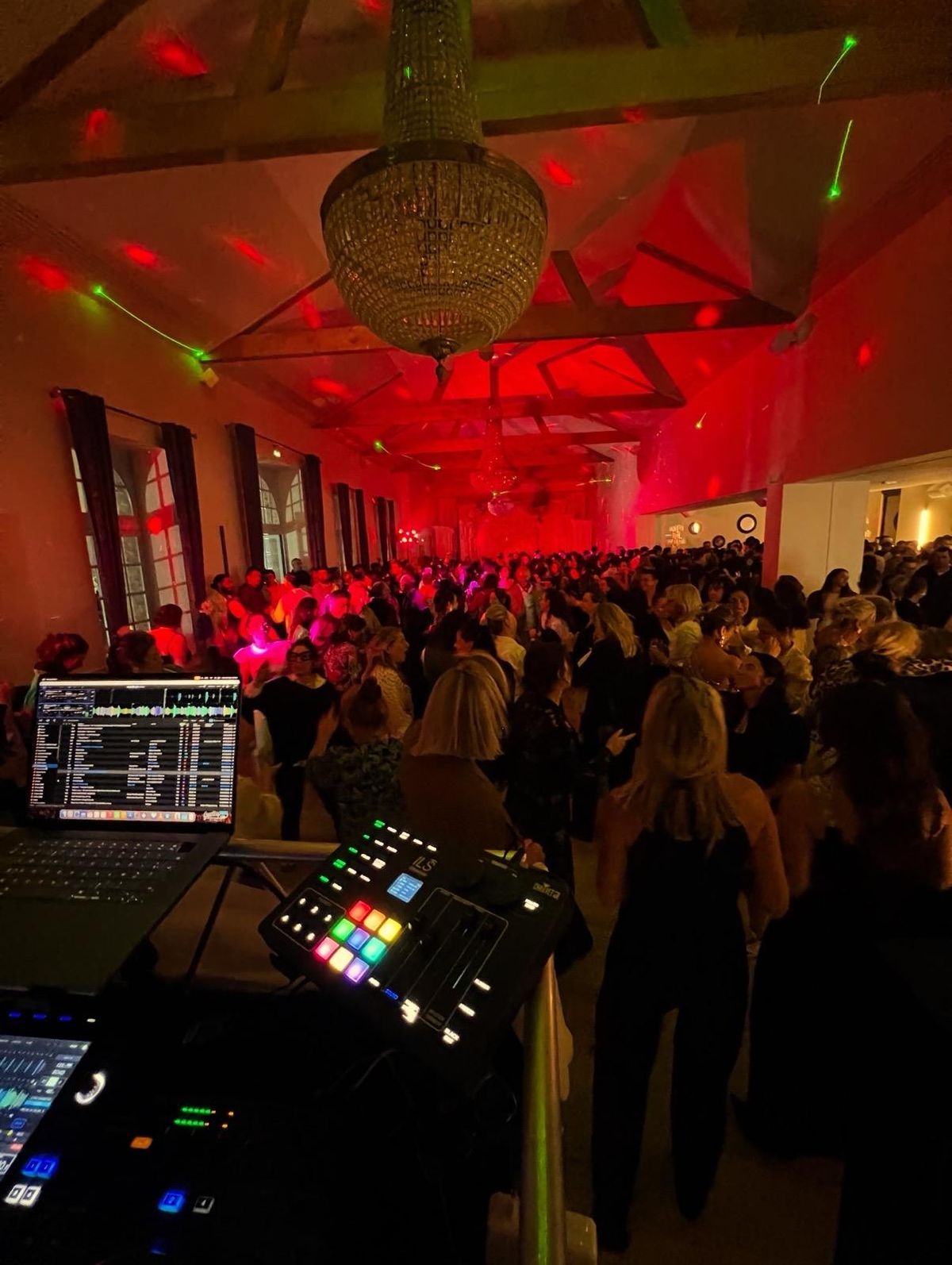A crowded dance floor at night under red lighting in a beamed reception hall, with a DJ booth in the foreground.