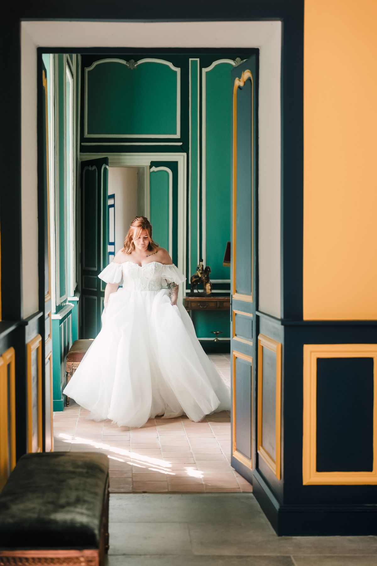 A bride in a ballgown walking through a doorway into a teal-panelled salon.