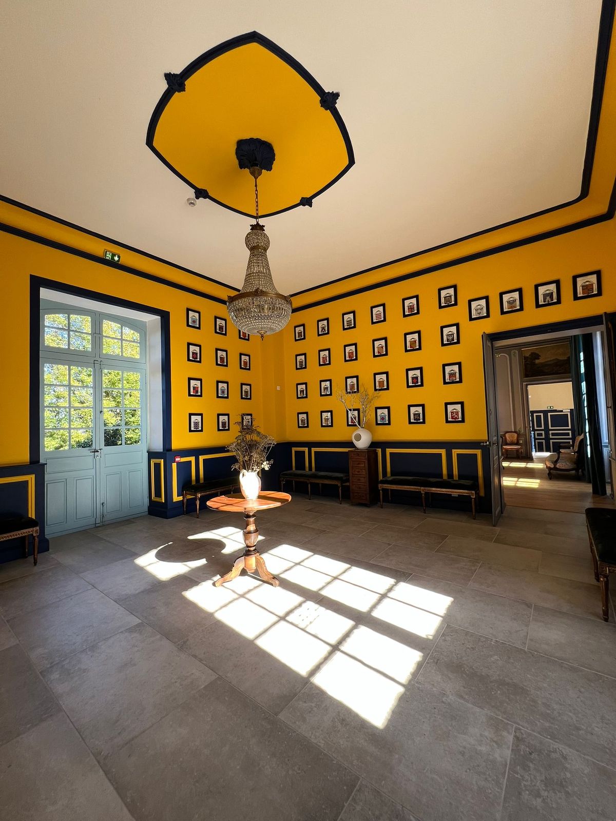 A formal yellow and black salon with a chandelier, framed portraits along the walls and period furniture.
