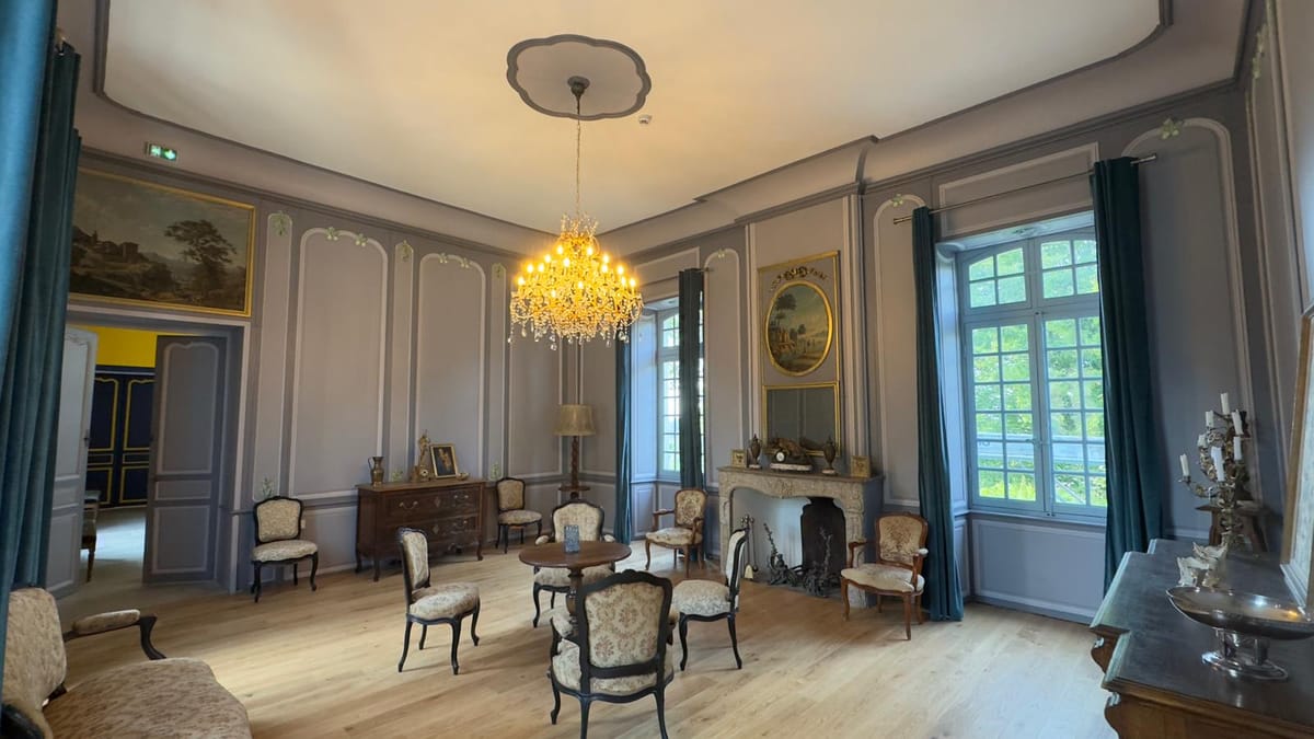 A blue-grey panelled salon with a stone fireplace, crystal chandelier and period armchairs.