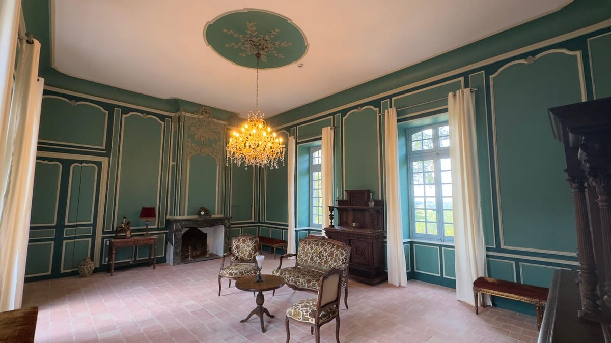 A teal-panelled salon with a stone fireplace, crystal chandelier and period armchairs arranged for conversation.
