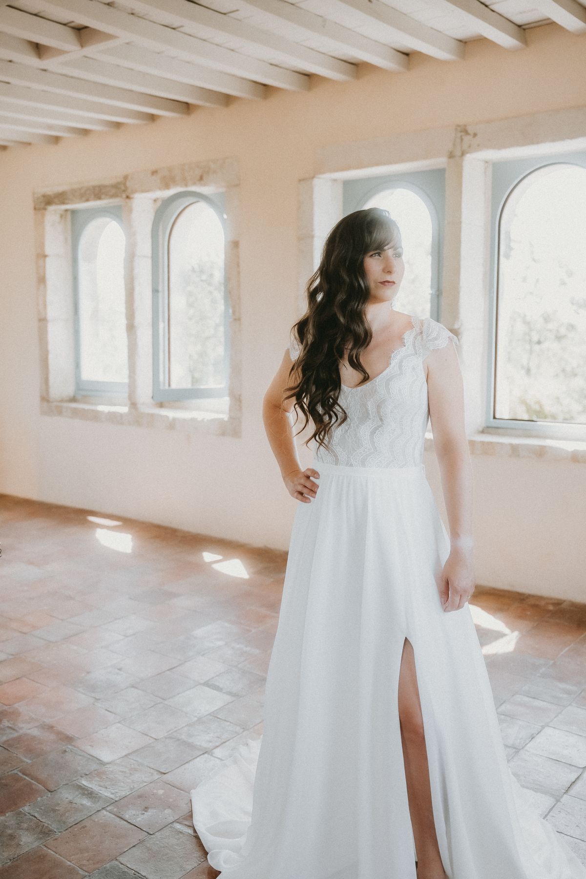 A bride in a white gown standing in a bright, whitewashed room with arched windows and a beamed ceiling.