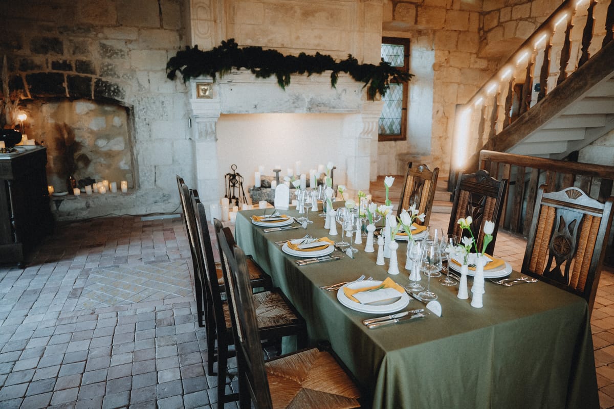 A formal dinner table set in a stone hall with a garland-draped fireplace mantel and a wooden staircase.