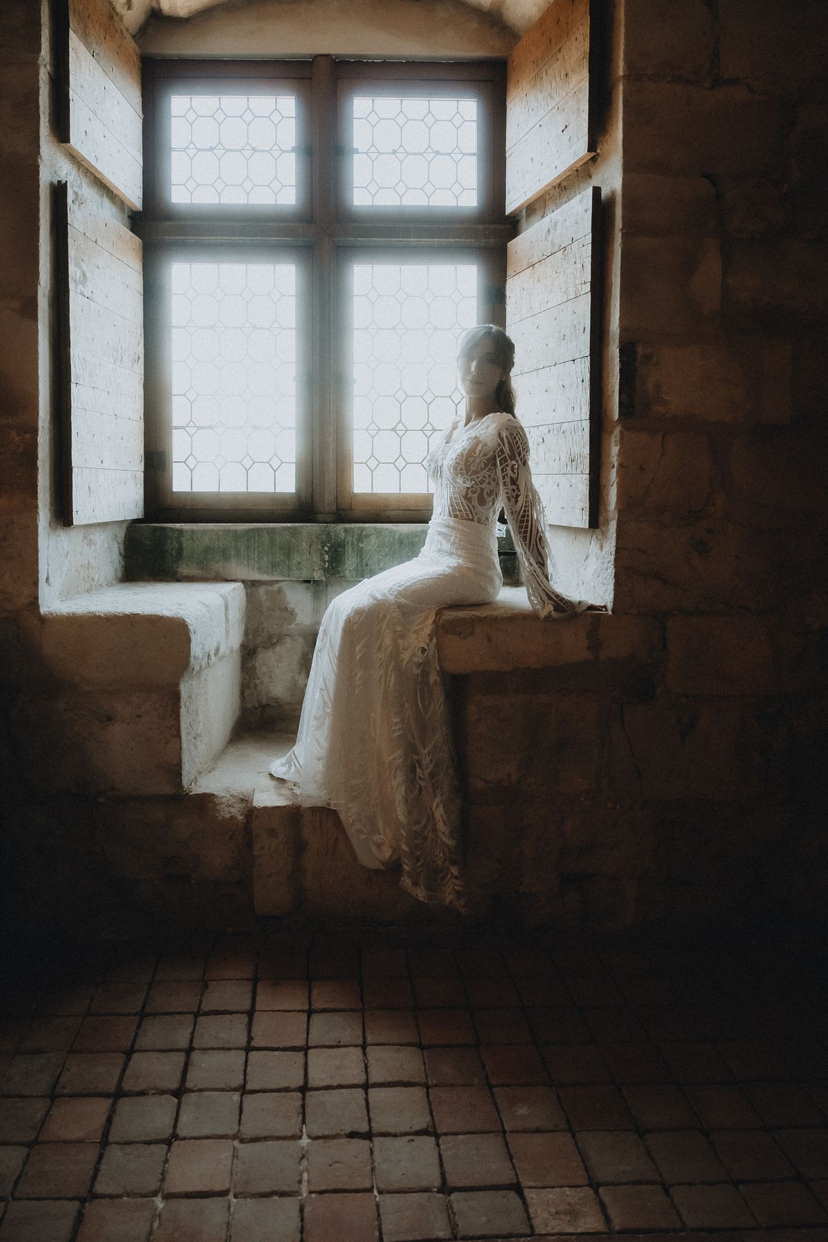 A bride in a lace gown seated in a stone window nook.