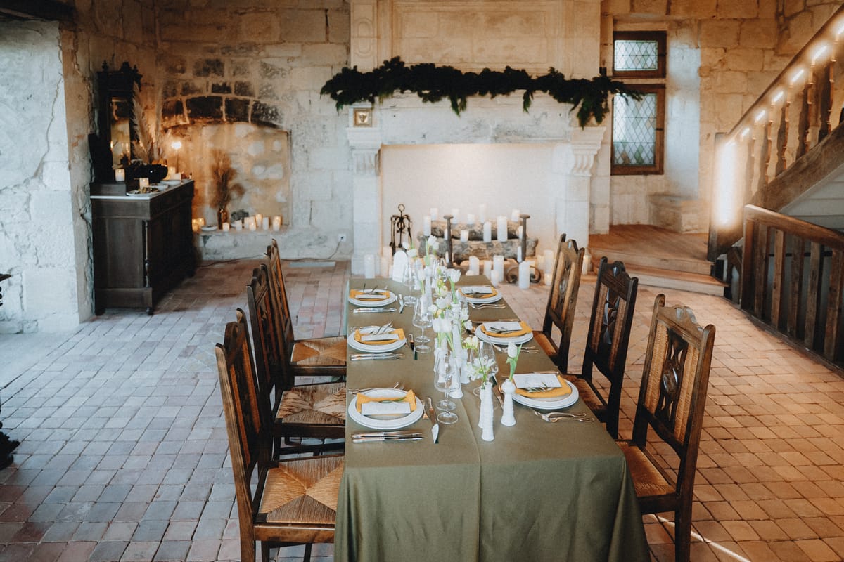A long dining table set for a winter dinner in a stone hall, with garland on the mantel and candlelight.