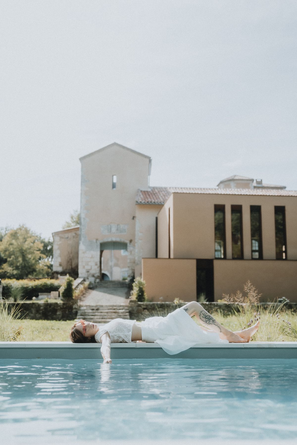 A bride in a white dress reclining at the edge of the swimming pool, with the château's modern extension building behind.