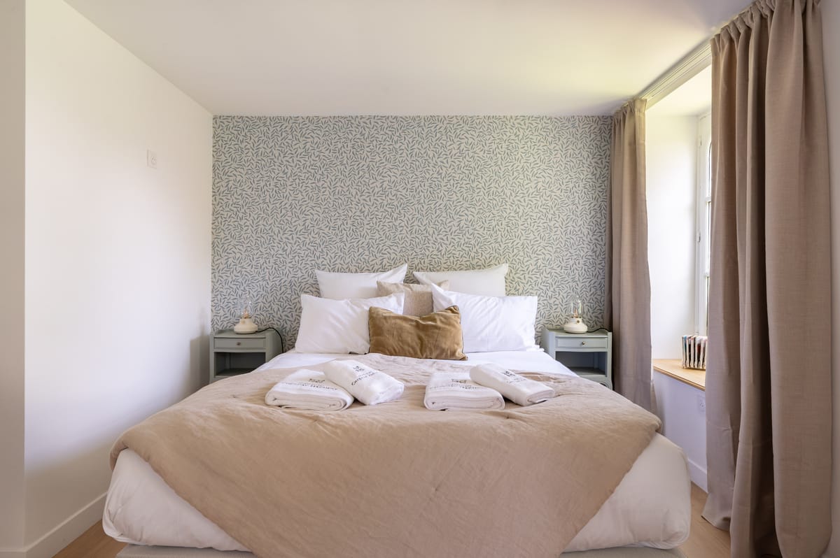 A double bedroom in the guest house with a patterned feature wall, linen bedding and folded towels