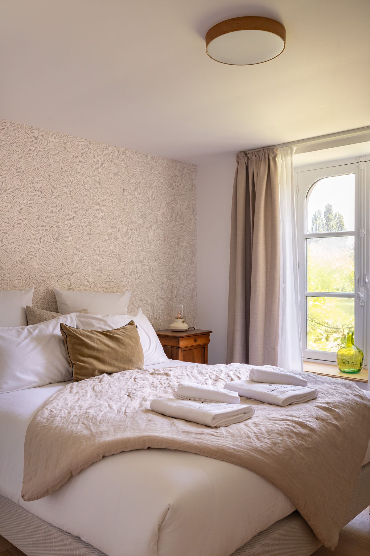 A guest bedroom with an arched window, wooden bedside table and layered white and linen bedding