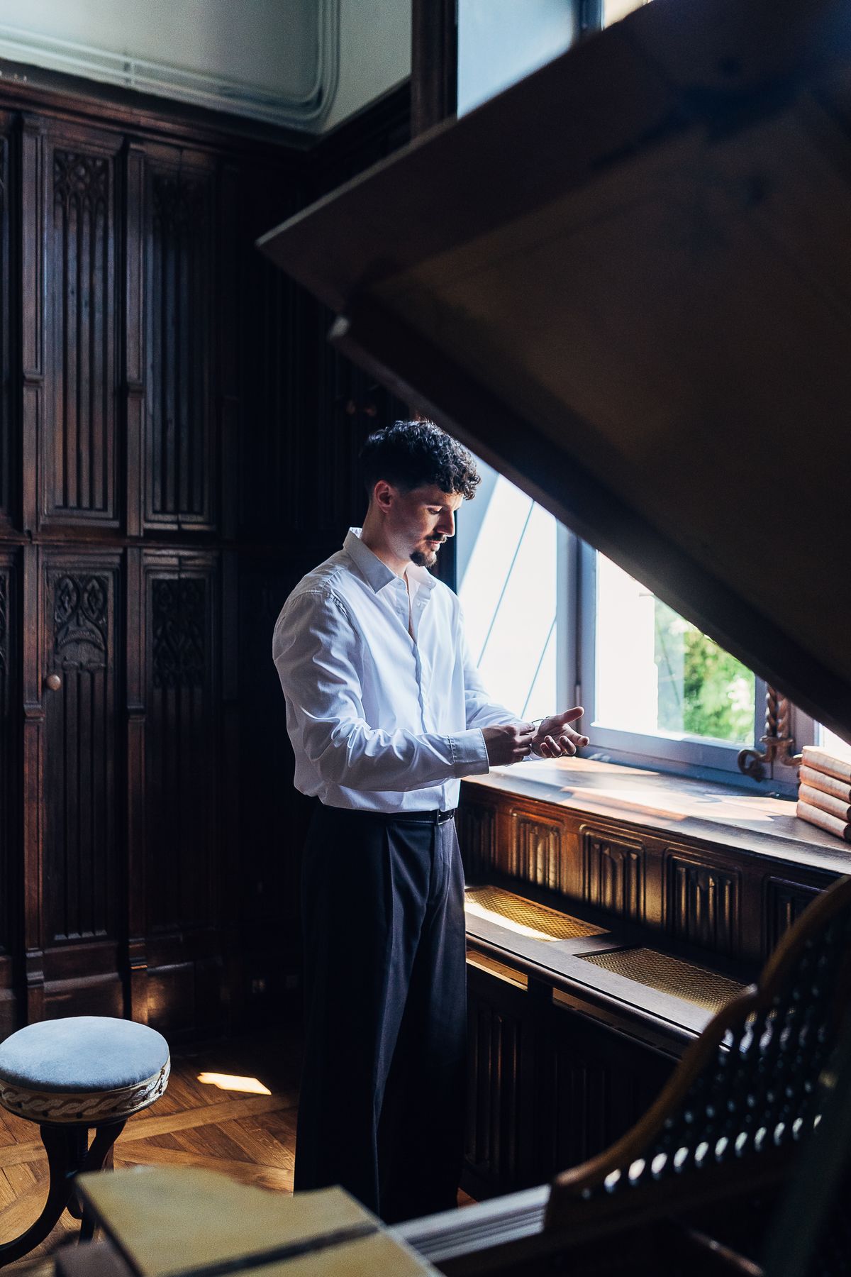 A groom fastening his cuff at a tall window of a château salon, with carved oak panelling and parquet