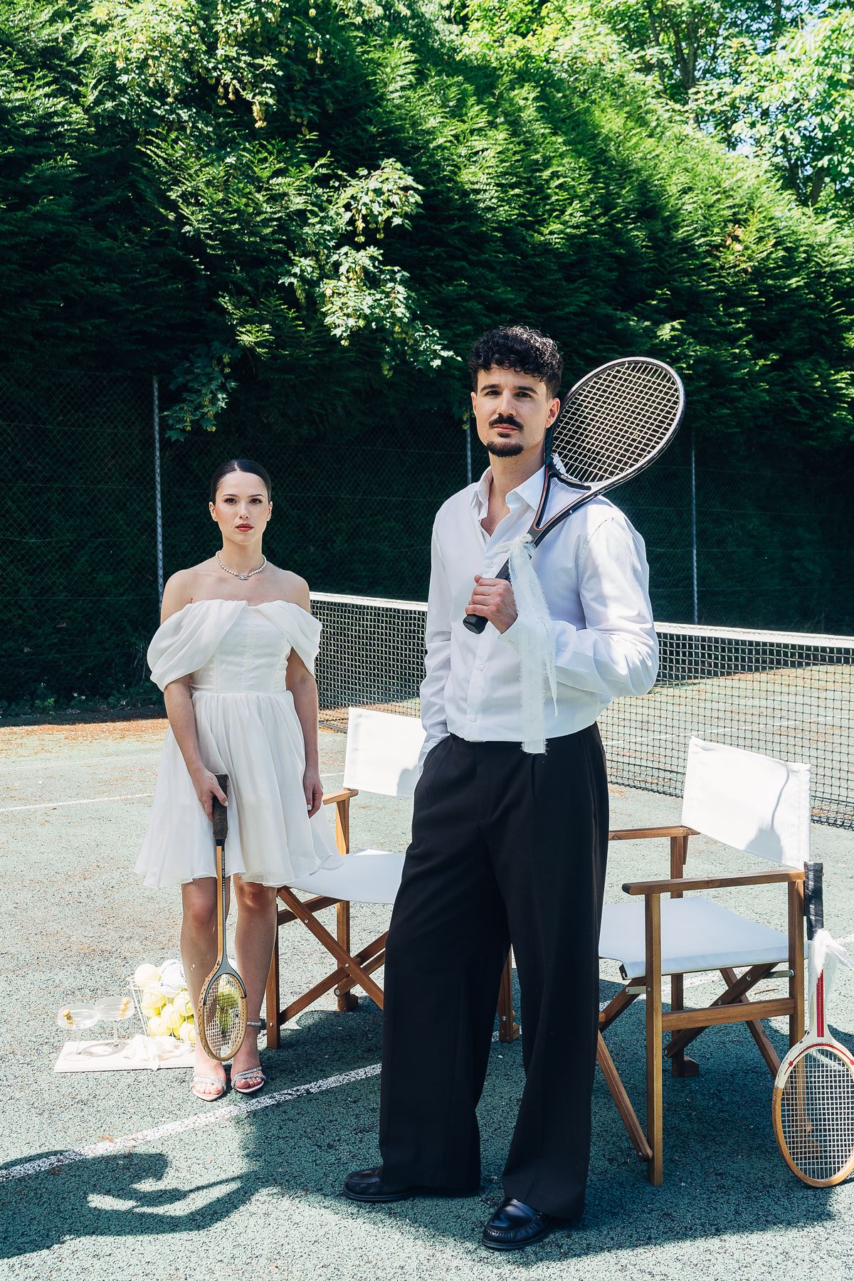 A bride and groom on the estate's tennis court with wooden racquets and directors' chairs, hedged by trees