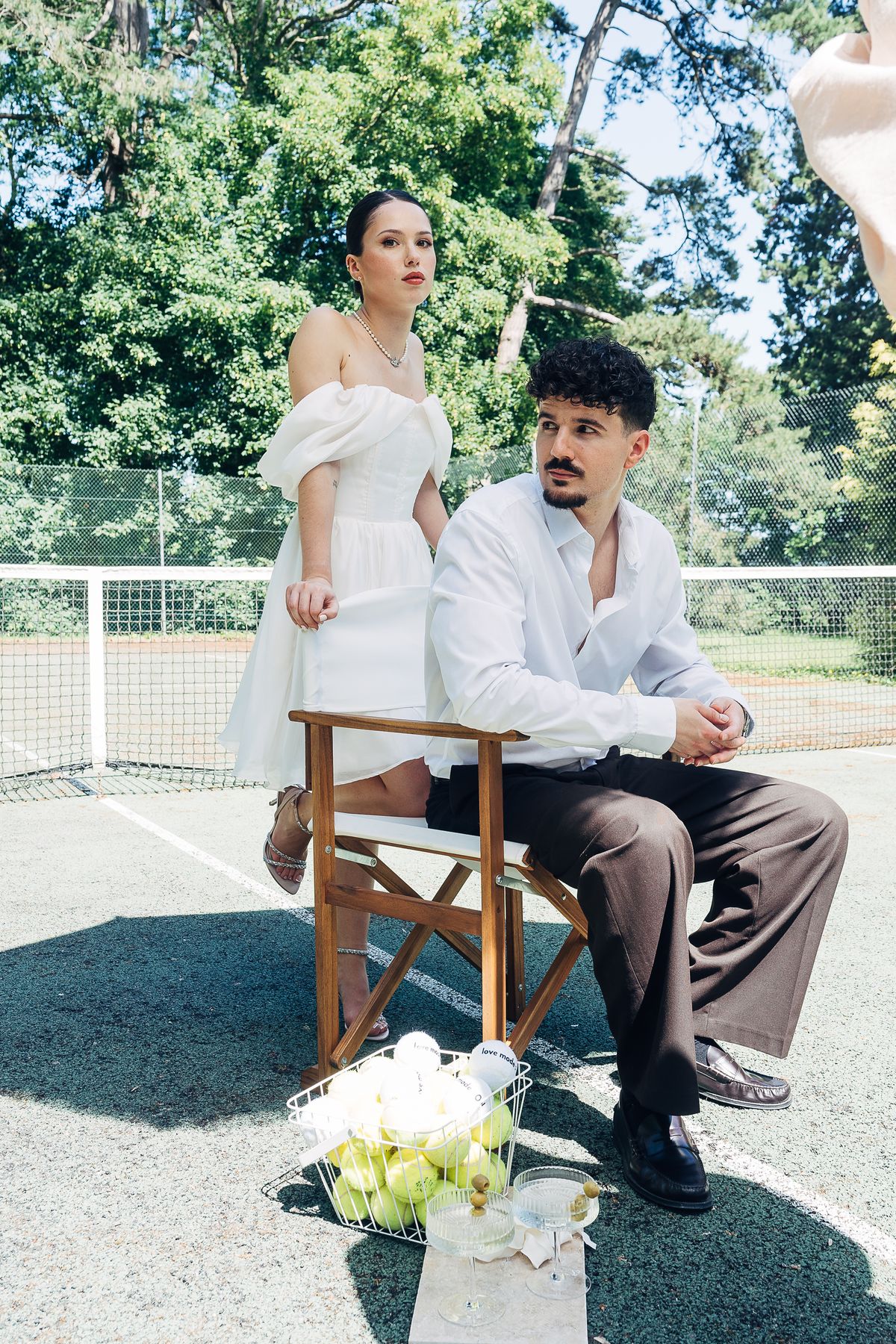 A groom seated in a directors' chair by the tennis net, the bride standing behind him, a basket of balls alongside