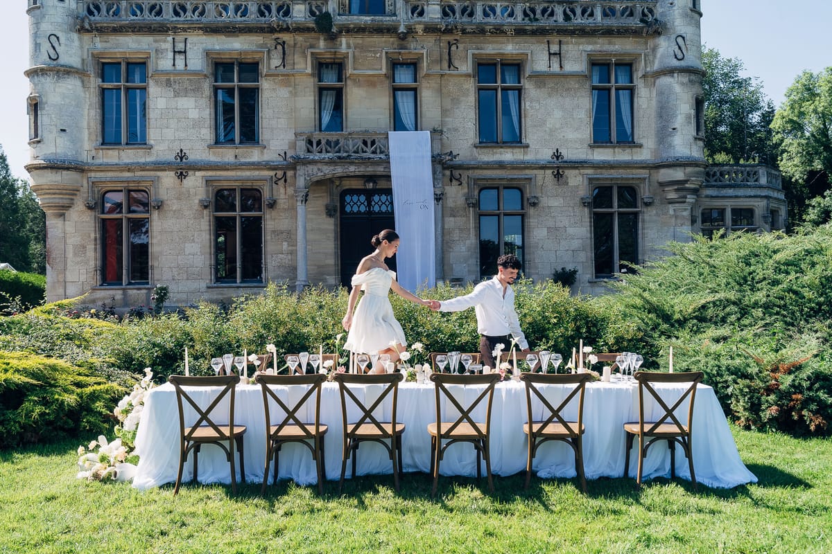 A bride and groom hand in hand behind a long table laid on the lawn below the château, with cross-back chairs