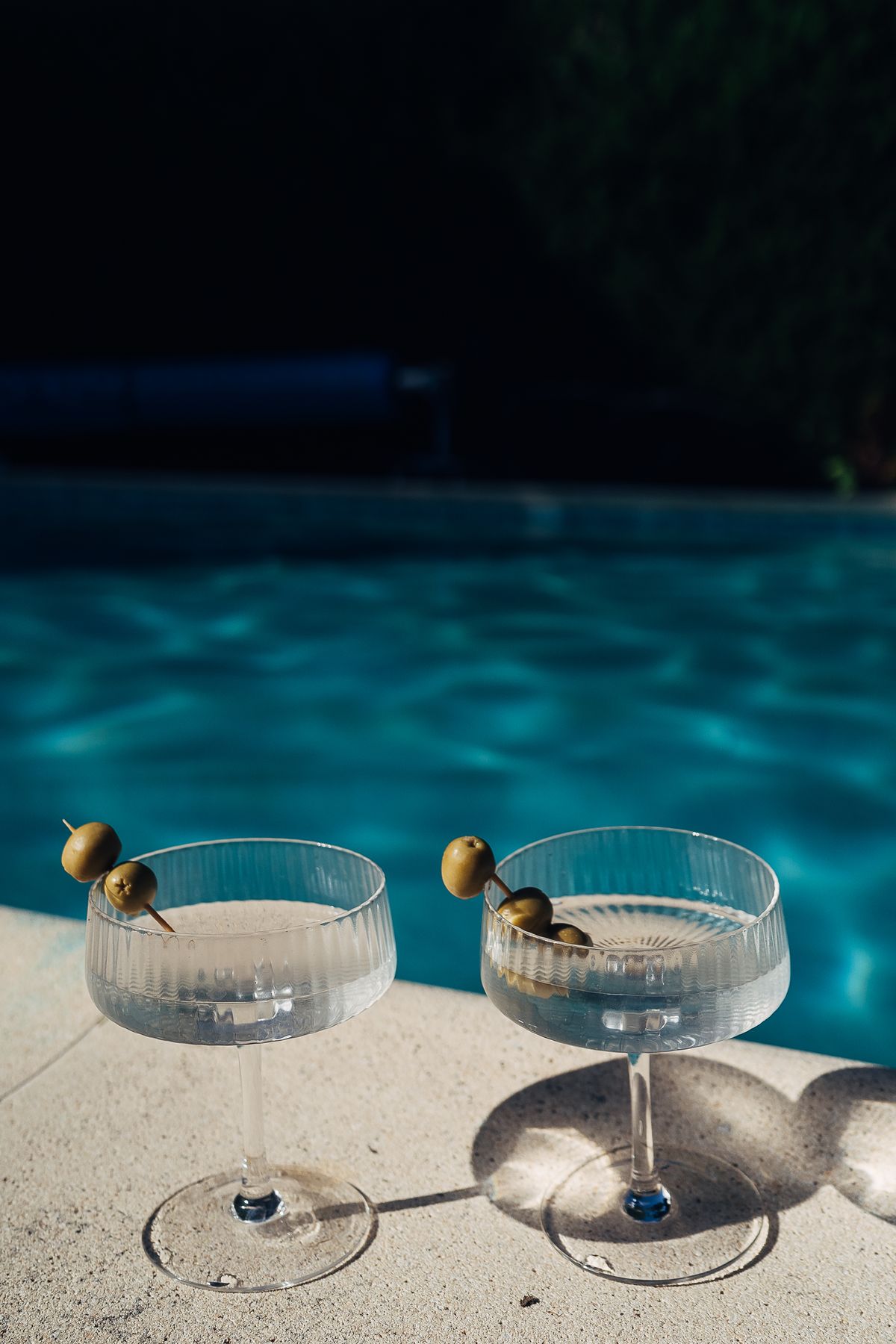 Two coupe glasses with olives on the poolside stone, the lit water behind them