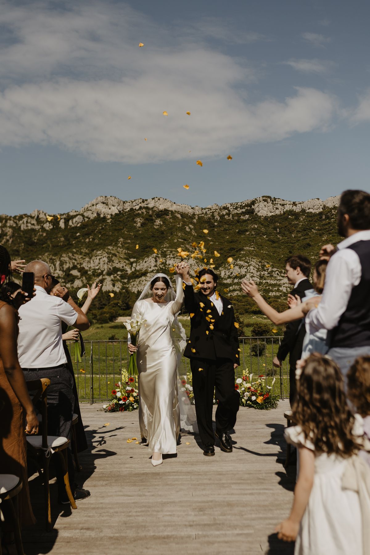Guests throw petals as a couple walk back down the aisle on the terrace at Domaine ODDO Vallon des Glauges, with the Alpilles behind