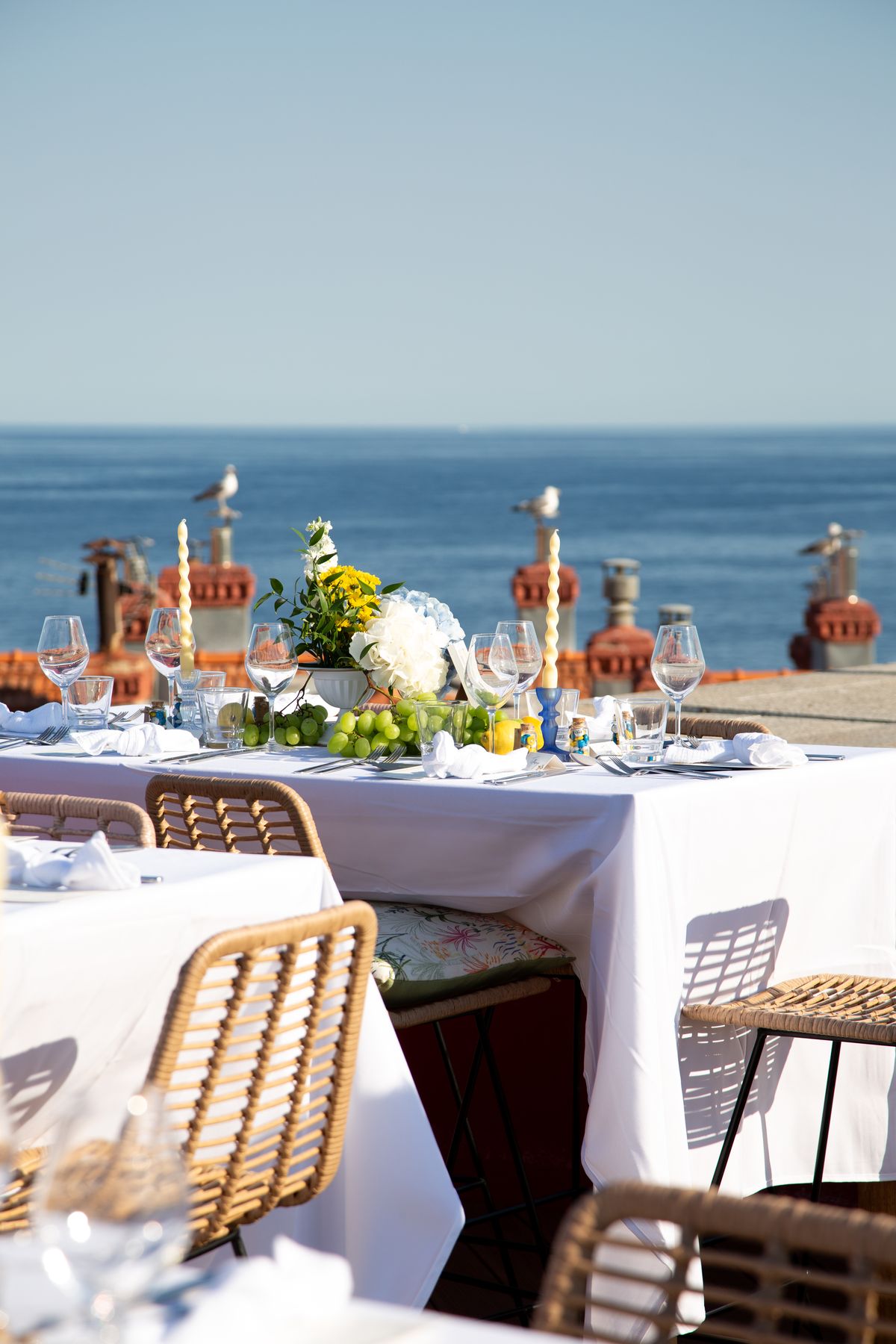 Dinner tables with white cloths and rattan chairs on the rooftop, with the Mediterranean behind