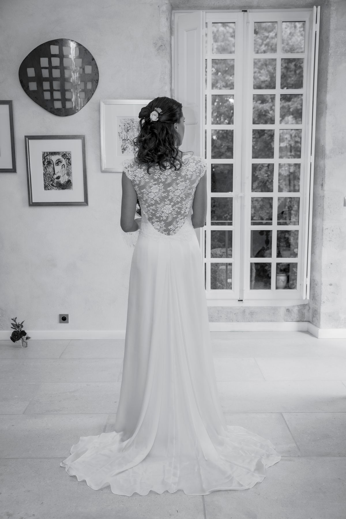 A bride at a tall French window in an interior room with artwork on the walls