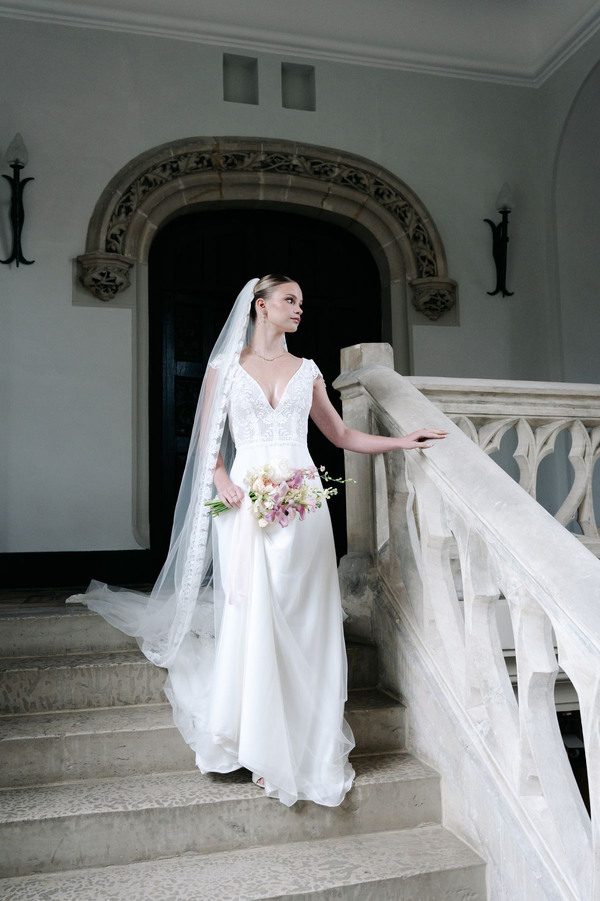 A bride in a lace gown and long veil descending the château's carved stone staircase, bouquet in hand