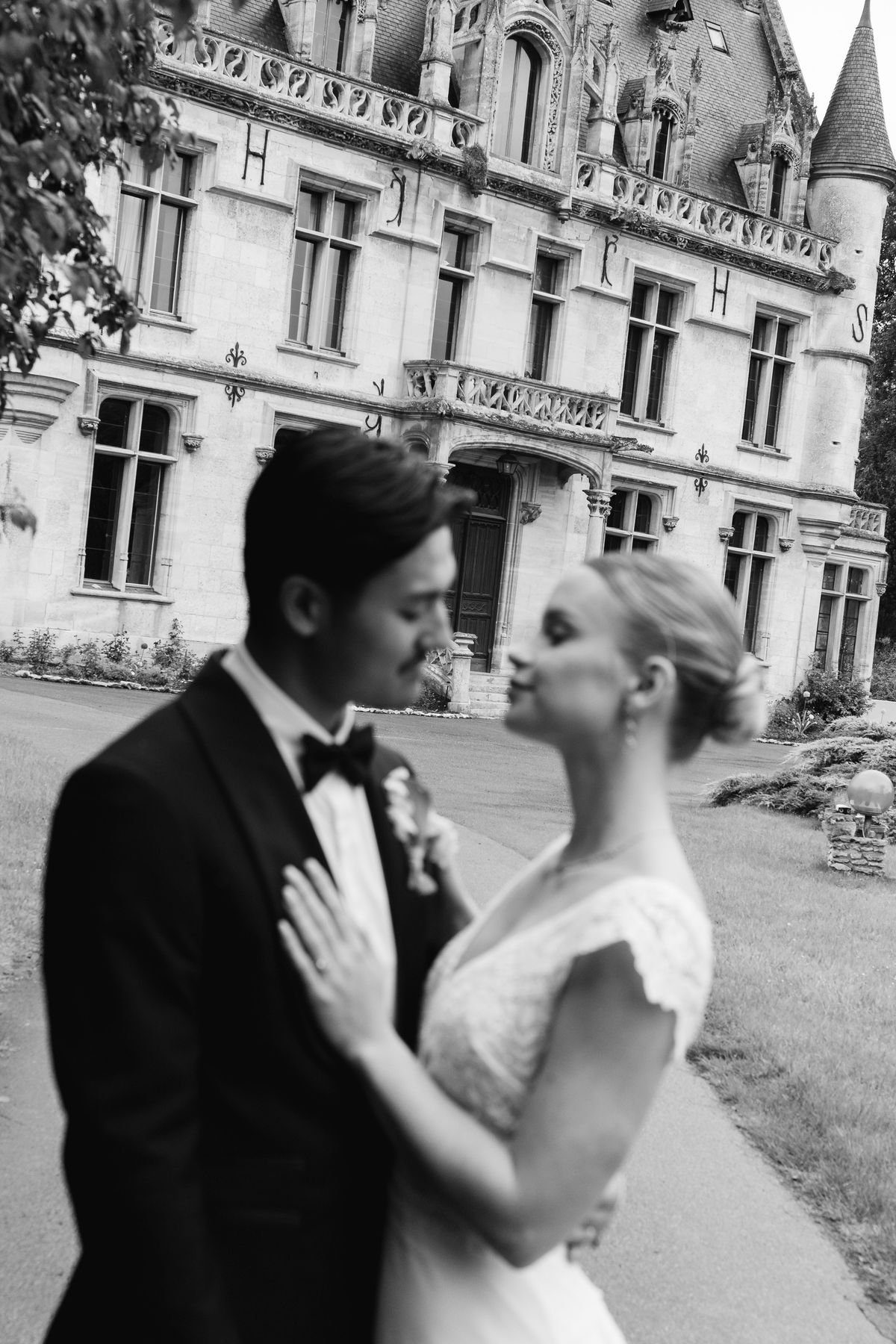 A bride and groom embracing with eyes closed in front of the château facade, in black and white