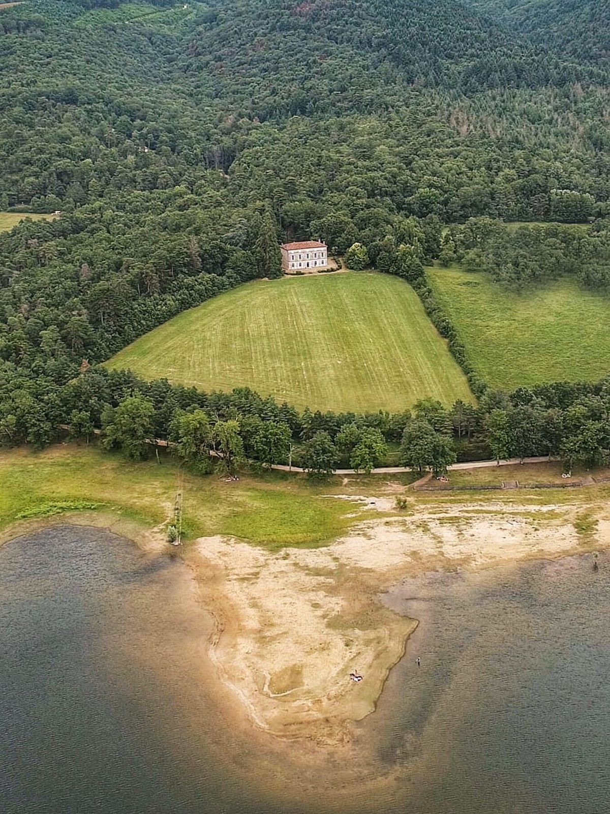 Aerial view of the château above its meadow, with a wooded shoreline and the sandy lake edge below