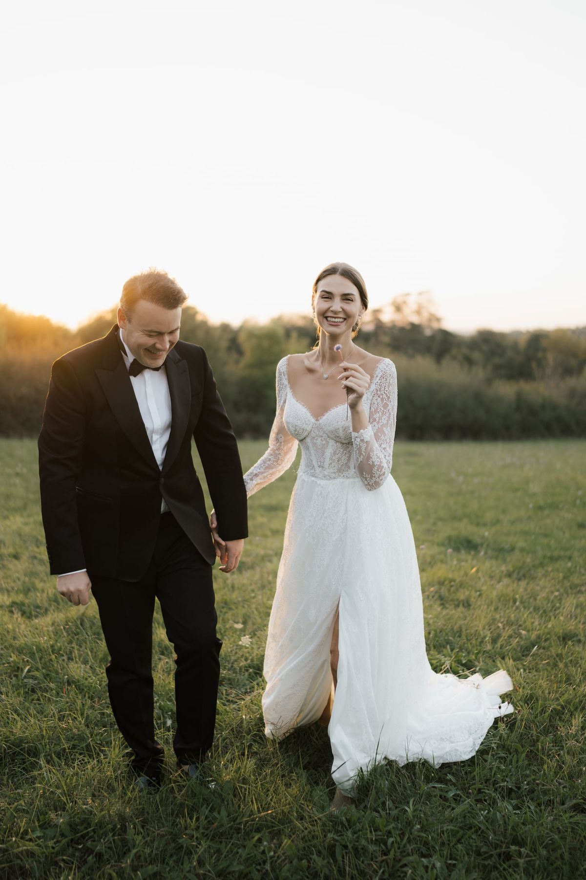 A bride and groom walking hand in hand through the meadow at golden hour