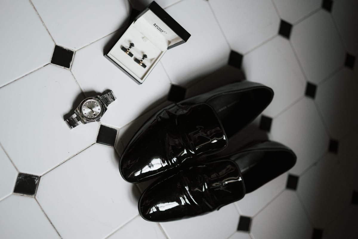 Patent shoes, a watch and cufflinks laid out on a black and white tiled floor