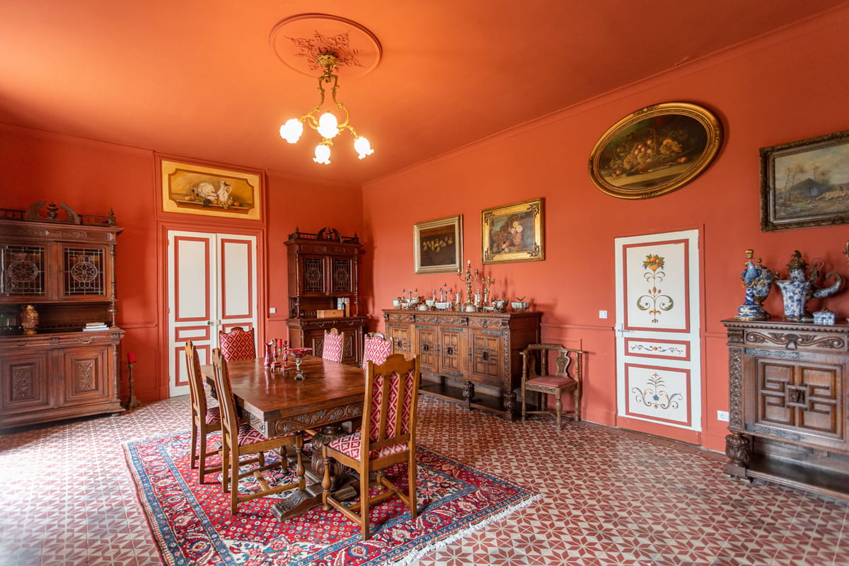 A red-walled room with a carved dining table and chairs on a rug, antique dressers and painted doors