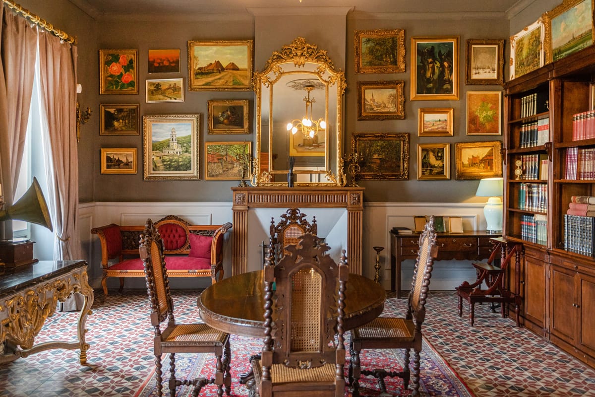 A salon hung with gilt-framed paintings around a gilded mirror, with bookshelves and a round table
