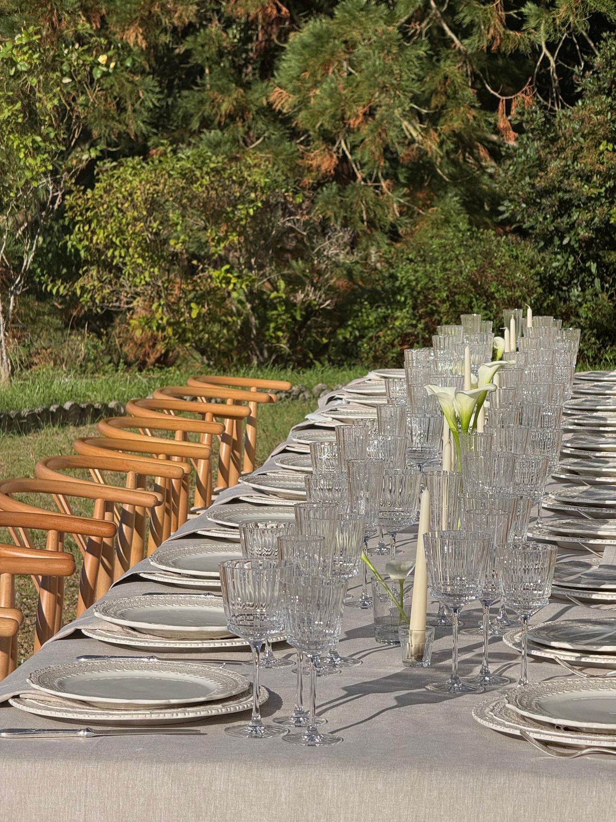 Crystal glassware and calla lilies along a long table set outdoors, with wooden chairs alongside
