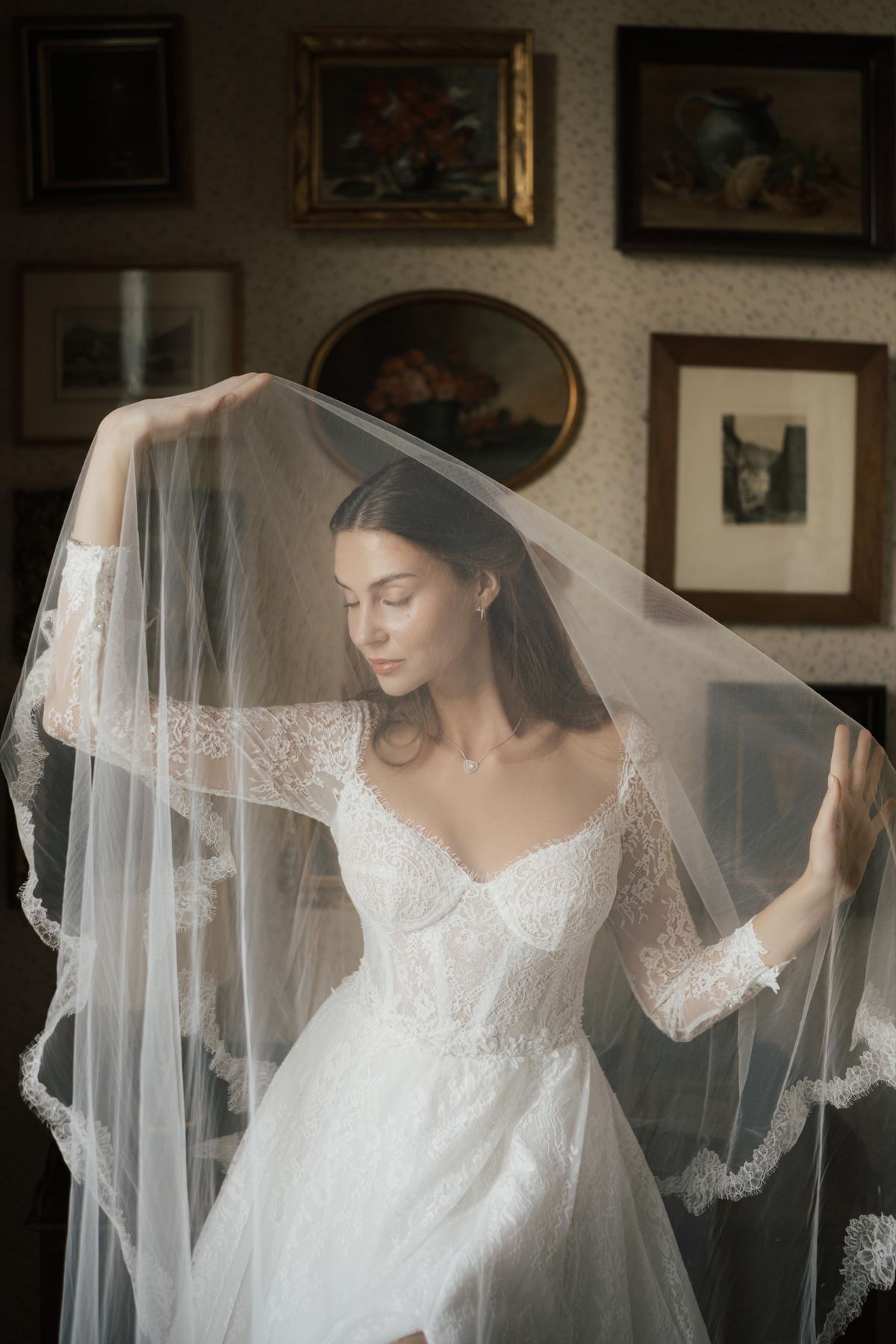 A bride lifting her veil in a light-filled room hung with framed artwork, getting ready on the wedding morning.
