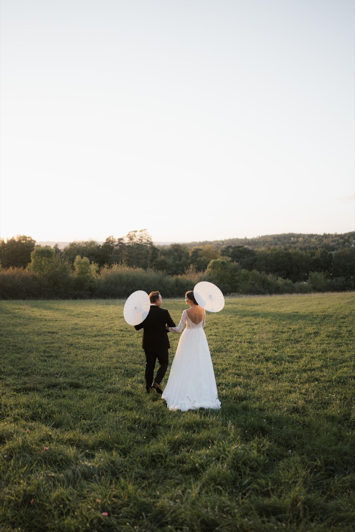 A couple walking hand in hand across the estate's meadow at sunset, carrying paper parasols.
