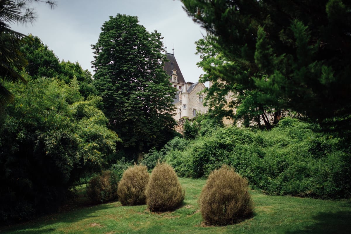 Clipped shrubs on a lawn below tall trees, with a château tower rising behind the greenery